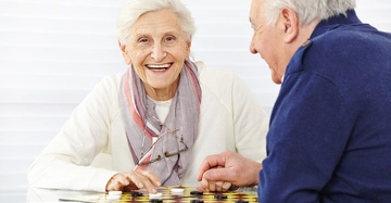 Ältere Frau mit Brille spielt Dame an einem Glastisch mit einem älteren Mann in einem hellen Raum. Sie lächelt, beide konzentrieren sich auf das Spiel.