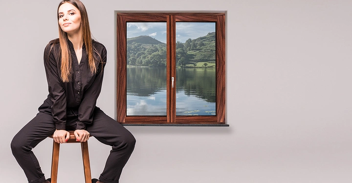 Eine Frau in schwarzer Kleidung sitzt auf einem Hocker in einem Raum mit hellem Hintergrund und einem Fenster, das eine Landschaft mit See und Bergen zeigt.