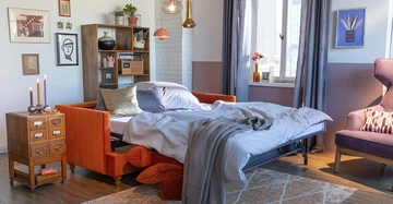 Ein ausgezogener Schlafsofa mit Bettwäsche im Wohnzimmer steht vor einem Bücherregal und neben einem Nachttisch mit Kerzen in einer gemütlich eingerichteten Wohnung mit Dekoration und violettem Sessel.