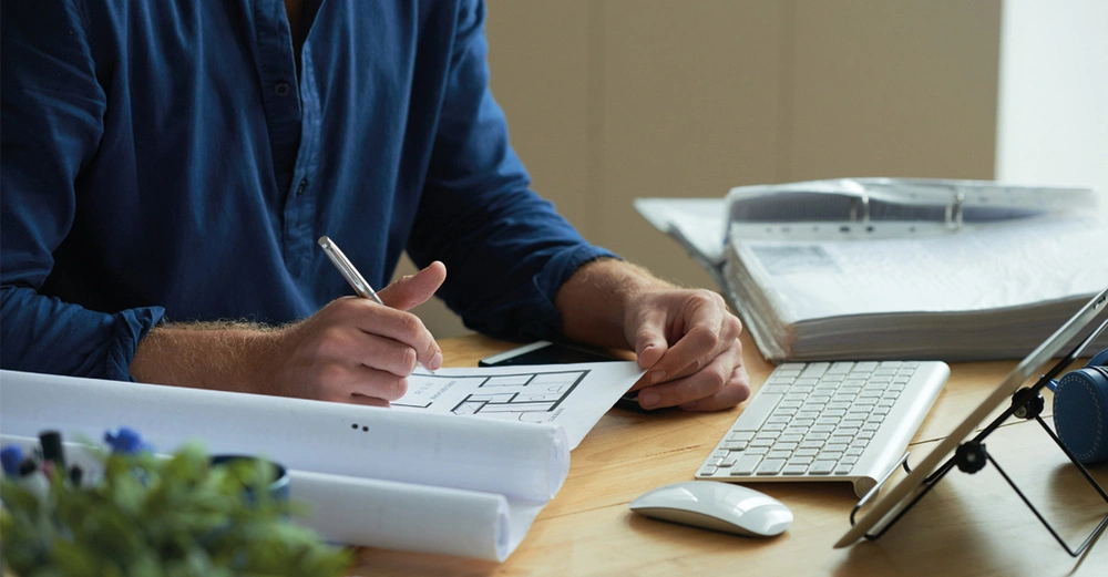 Mann in blauem Hemd arbeitet am Schreibtisch sitzend an einem Bauplan, auf dem Tisch befinden sich eine dicke Dokumentenmappe und ein Tablet samt Maus und Tastatur