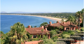 Panoramablick auf eine weitläufige Küstenbucht mit türkisblauem Meer, langem Sandstrand und mediterranen Villen mit Palmen im Vordergrund unter klarem Himmel.