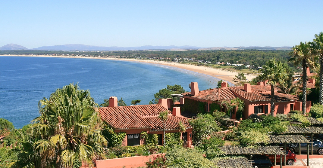 Panoramablick auf eine weitläufige Küstenbucht mit türkisblauem Meer, langem Sandstrand und mediterranen Villen mit Palmen im Vordergrund unter klarem Himmel.