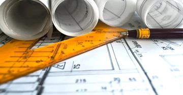 Mehrere zusammengerollte Baupläne liegen auf einem Tisch mit einem Lineal und einem Kugelschreiber darauf verstreut. Die Baupläne zeigen technische Zeichnungen und Diagramme im Büro eines Architekten.