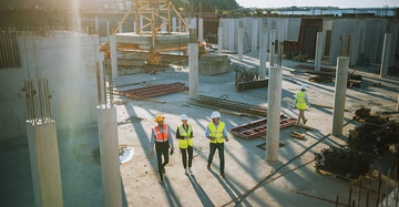Arbeiter gehen über eine Baustelle zwischen Betonpfeilern und Baumaterialien bei Tageslicht. Sie tragen Schutzhelme und Warnwesten während sie den Platz inspizieren. Im Hintergrund sind weitere Baustellenstrukturen.