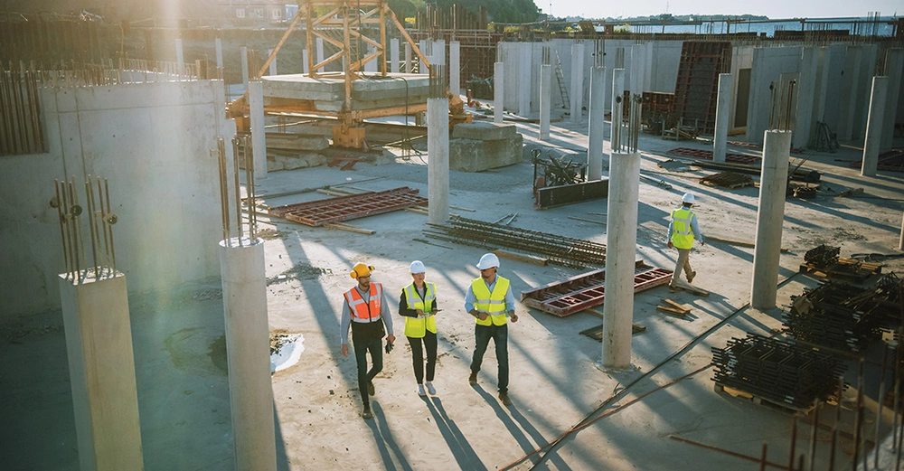 Arbeiter gehen über eine Baustelle zwischen Betonpfeilern und Baumaterialien bei Tageslicht. Sie tragen Schutzhelme und Warnwesten während sie den Platz inspizieren. Im Hintergrund sind weitere Baustellenstrukturen.