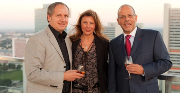 Galerie-Bild 36: Drei Personen stehen mit Weingläsern in der Hand auf einer Terrasse im Freien. Im Hintergrund sind Hochhäuser und eine Stadtlandschaft zu sehen. Alle Personen tragen formelle Kleidung.