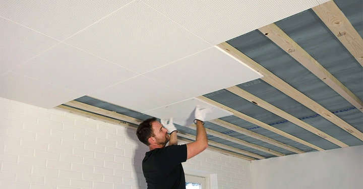 Galerie-Bild 3: Ein Mann installiert Deckenplatten in einem Raum mit weiß gestrichenen Ziegelwänden und einem Fenster im Hintergrund.