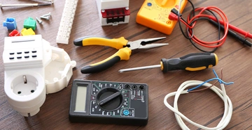 Verschiedene Elektrowerkzeuge und -komponenten liegen auf einem Holztisch; darunter ein Multimeter, eine Zange, ein Schraubendreher, Kabel, Isolierklemmen und bunte Klemmen.