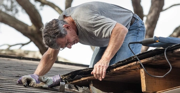 Mann in grauem T-Shirt und Jeans arbeitet auf einem Hausdach mit Handschuhen und einem Elektrowerkzeug, um Dachbeschädigungen zu reparieren Bäume im Hintergrund