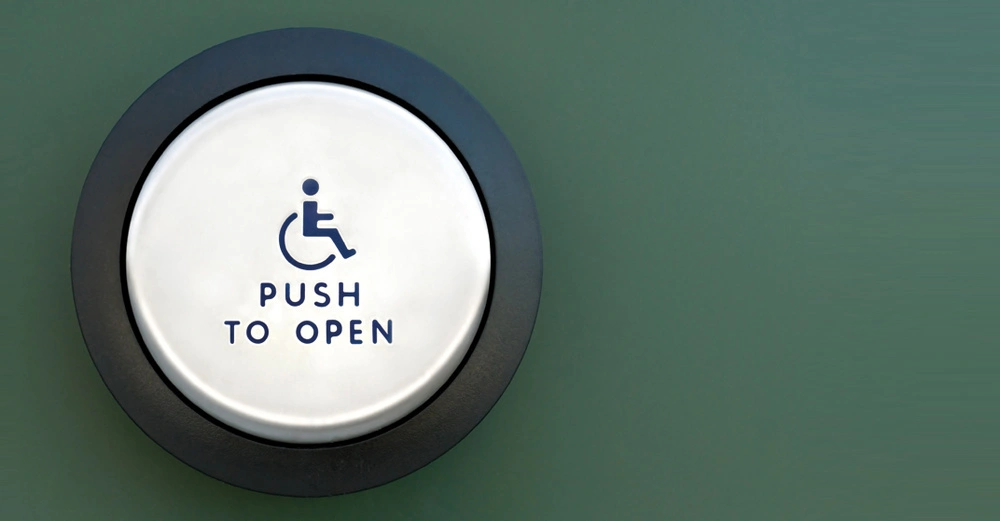 weißer runder Schalter mit schwarzem Rahmen, darauf ein Rollstuhlfahrer und die Worte "Push to open". Die Wand, auf der der Schalter sich befindet ist moosgrün. 
