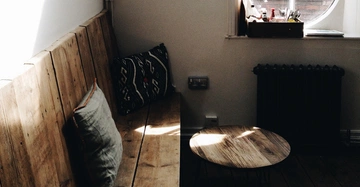 Holzbank mit Kissen steht an einer weißen Wand in einem Zimmer neben einem kleinen runden Tisch aus Holz. Ein Fenster und ein Heizkörper befinden sich im Hintergrund.