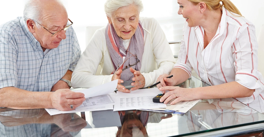 Zwei ältere Personen sitzen an einem Tisch und betrachten Dokumente während die jüngere Person mit einem Stift in der Hand auf die Dokumente zeigt. Hintergrund ist ein heller Raum.