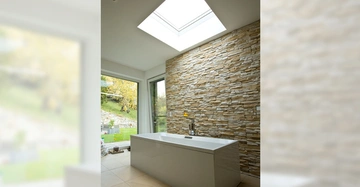 Weiße Badewanne stehend vor einer Wand mit Steinverkleidung in einem modernen Badezimmer mit großem Oberlicht und Blick auf einen grünen Garten durch bodentiefe Fenster.