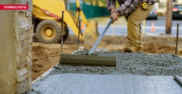 Ein Bauarbeiter glättet frischen Beton mit einem Werkzeug Baustelle im Freien mit gelbem Bagger im Hintergrund Mauer auf der linken Seite Text SYMBOLFOTO in der oberen linken Ecke