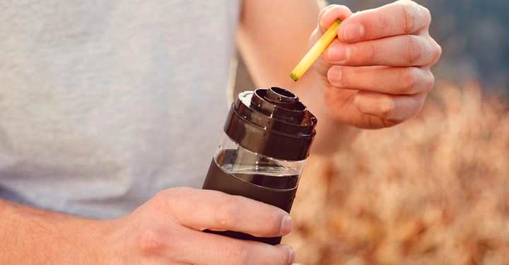 Zwei Hände halten eine schwarze Thermosflasche und ein kleines gelbes Objekt auf einer sonnigen, natürlichen Außenseite.