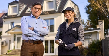 Mann in Hemd und Hose posiert mit junger Handwerkerin vor einem Haus