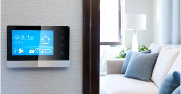 Smart Home Panel an der Wand zeigt Datum, Temperatur, Akkuladung und weitere Features. Im Hintergrund Couch mit Zierkissen.