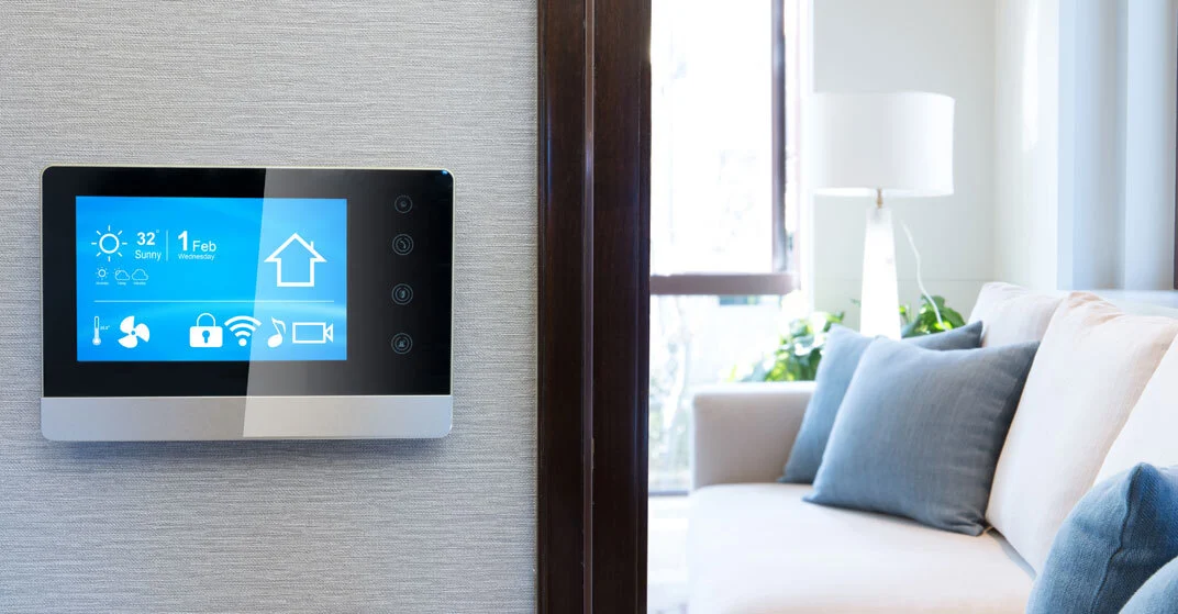 Smart Home Panel an der Wand zeigt Datum, Temperatur, Akkuladung und weitere Features. Im Hintergrund Couch mit Zierkissen.
