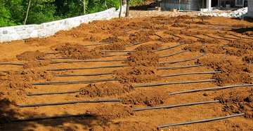 Rohrleitungen verlaufen parallel über einen frisch umgegrabenen, erdigen Gartenbereich neben einer teilweise fertiggestellten Hauskonstruktion; Bäume und eine Steinmauer umgeben das Grundstück.