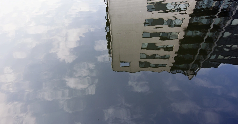 Spiegelbild eines mehrstöckigen Gebäudes mit Fenstern in ruhigem Wasser Himmel und Wolken sind ebenfalls reflektiert Keine weiteren Objekte oder Schriftelemente im Bild zu sehen