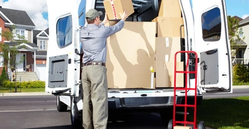 Ein Mann lädt Kartons in einen Lieferwagen vor einem modernen Wohnhaus ein. Neben ihm steht eine rote Sackkarre mit einem weiteren Karton.