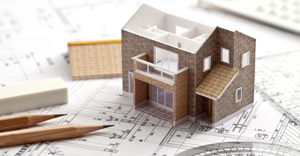 Ein Modellhaus steht auf architektonischen Bauplänen mit zwei Bleistiften und einem Radiergummi daneben im Hintergrund sind Lineale und ein Taschenrechner zu sehen.