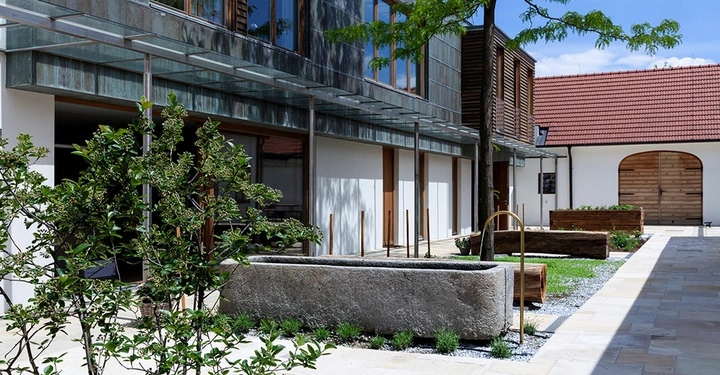 Galerie-Bild 1: Steingefäß steht still in einem modernen Innenhof mit Glasdach und Pflanzen umgeben von Gebäuden mit hölzernen und verglasten Fassaden.