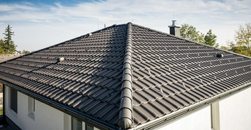 Dachziegel sind auf einem großen geneigten Dach angebracht umgeben von Bäumen und blauem Himmel in einem Wohngebiet.