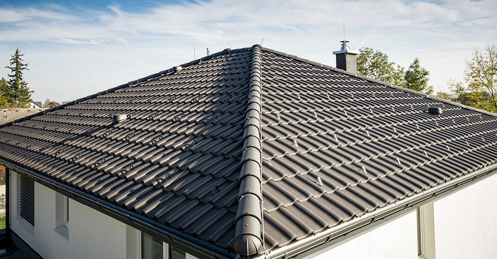 Dachziegel sind auf einem großen geneigten Dach angebracht umgeben von Bäumen und blauem Himmel in einem Wohngebiet.