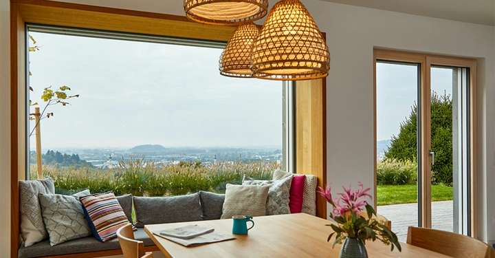 Holztisch mit Blumen und Tassen steht in einem gemütlichen Essbereich vor großen Fenstern Baumuster und Sitzkissen erhellen den Raum mit Blick auf eine grüne Landschaft und die Ferne