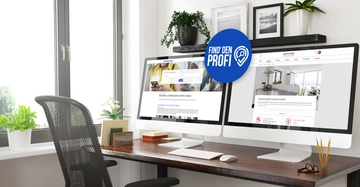 Zwei Computerbildschirme auf einem Schreibtisch zeigen Websites an; Find' den Profi steht auf einem blauen Symbol. Im Hintergrund Pflanzen und Bilder in einem modernen Büro mit großem Fenster.