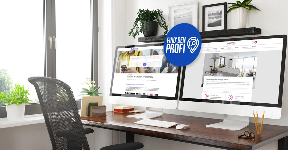 Zwei Computerbildschirme auf einem Schreibtisch zeigen Websites an; Find' den Profi steht auf einem blauen Symbol. Im Hintergrund Pflanzen und Bilder in einem modernen Büro mit großem Fenster.