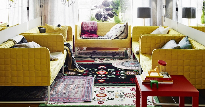 Gelbe Sofas stehen in einem gemütlichen Wohnzimmer reichlich dekoriert mit bunten Teppichen und Kissen ein roter Couchtisch mit Büchern und einem Dekorobjekt im Vordergrund graue Lampe und Pflanzen im Hintergrund.