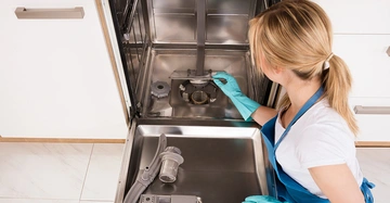 blonde junge Frau hockt auf Boden und wischt mit blauen Putzhandschuhen und Schürze bekleidet den leeren Geschirrspüler aus