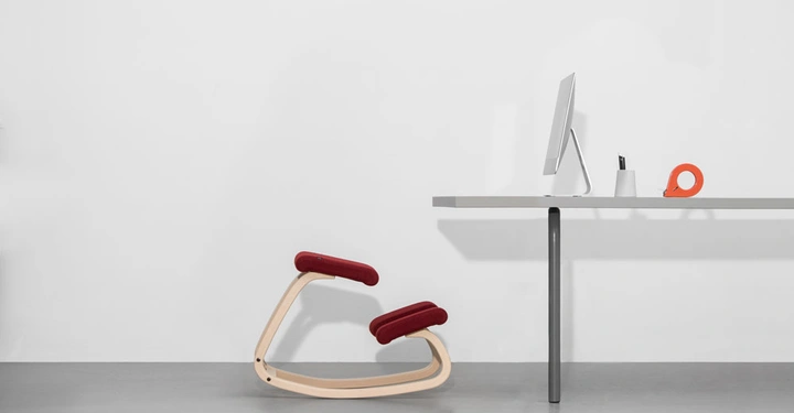 Ein ergonomischer Stuhl mit roter Polsterung steht auf dem Betonboden neben einem modernen Schreibtisch mit einem Computer und Büromaterialien in einem minimalistischen Raum.