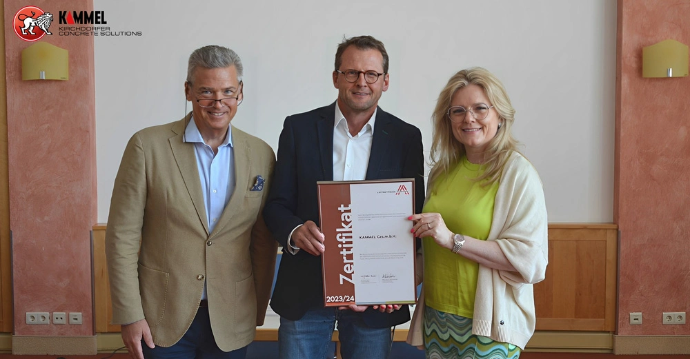 Drei Personen posieren gemeinsam in einem Konferenzraum während die mittlere Person ein Zertifikat hält. Links oben steht der Text KAMMEL KIRCHDORFER CONCRETE SOLUTIONS. Das Zertifikat trägt den Text: Zertifikat 2023/24 KAMMEL GmbH