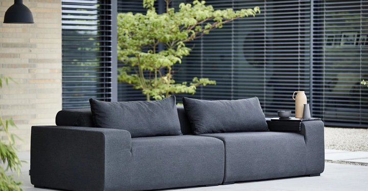 Galerie-Bild 2: Ein graues Sofa steht im Freien neben modernen Gebäuden und einem Baum im Hintergrund; darauf stehen eine beige Kanne und eine schwarze Tasse auf einem Tablett.