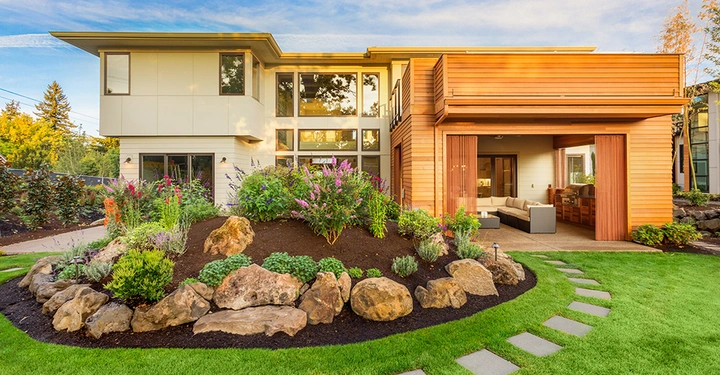 Modernes zweistöckiges Haus eine integrierte Veranda mit Sitzbereich daneben runde Gartenanlage mit Steinen und Pflanzen bei Sonnenuntergang umgeben von gepflegtem Rasen und Bäumen im Hintergrund