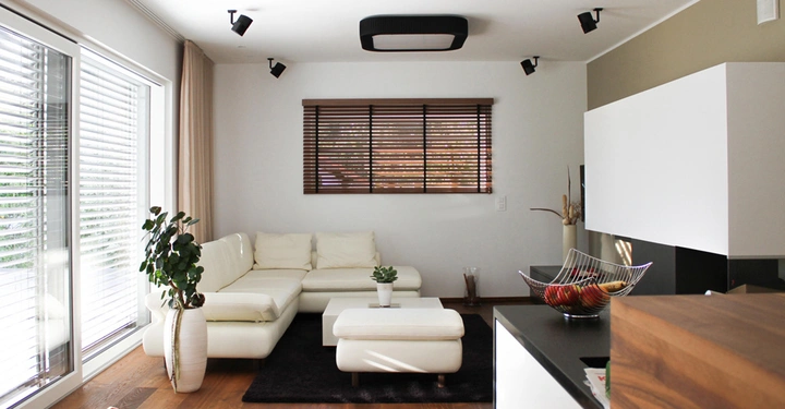 Weißes Ecksofa steht im Wohnzimmer vor einem Fenster mit Jalousien Pflanzen und Obstschale auf einem Couchtisch auf Holzfußboden in einem modernen Raum mit großen Glasfenstern.