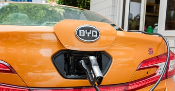 Ein orangefarbenes Auto lädt an einer Ladestation auf in einer häuslichen Umgebung mit Bäumen und einem weißen Gebäude im Hintergrund. Text BYD