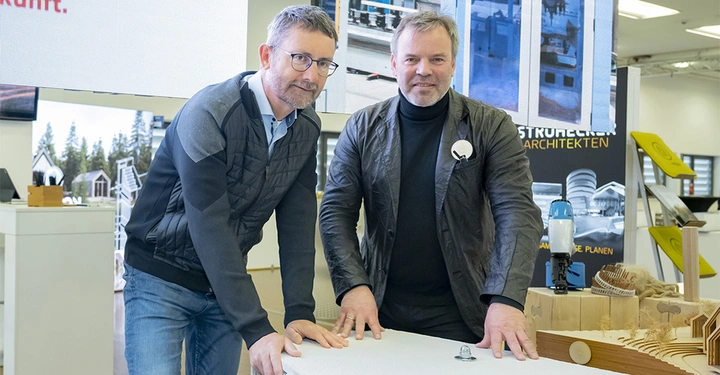 Zwei Männer beugen sich über ein weißes Modell in einem modernen Ausstellungraum mit Architekturmodellen und Plakaten im Hintergrund.