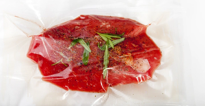 Ein vakuumverpacktes rohes Steak mit Gewürzen und frischen Kräutern, liegend auf einer weißen Oberfläche.
