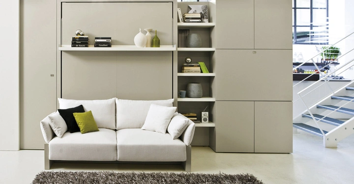 Ein beiges Sofa mit hellen und dunklen Kissen steht vor einem grauen Regal und Schrankwand in einem modernen Raum neben einer Treppe. Ein flauschiger Teppich liegt davor. Bücher auf einem Regal - "Stories by Jack London" - "Sons and Lovers by D.H. Lawrence" - "Casabella"