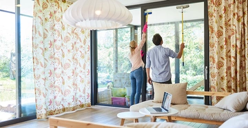 Zwei Personen reinigen gemeinsam große Glasfenster in einem sonnigen Wohnzimmer mit blumengemusterten Vorhängen, einem Holzsofa und einem Laptop auf dem Couchtisch.