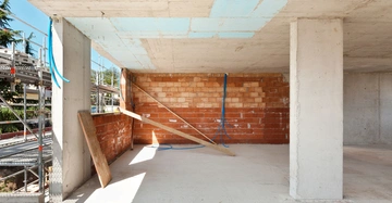 Unvollendete Betonstruktur mit zwei Betonstützen Wände aus roten Ziegelsteinen und blauem Elektrikkabel Baumaterialien stützend an der Wand Außenumgebung zeigt Gerüste und Vegetation unter klarem Himmel.