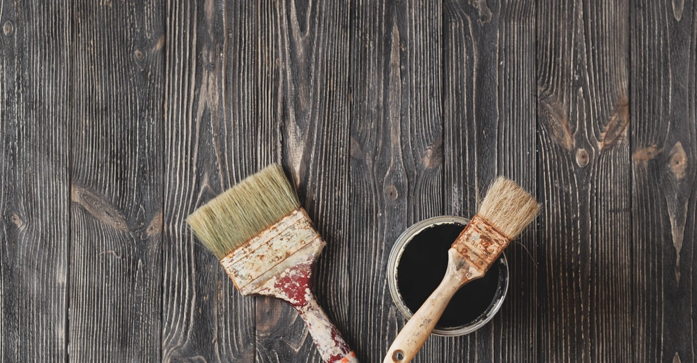 Zwei gebrauchte Pinsel liegen auf einem dunklen Holztisch Einer der Pinsel ist in eine Dose mit schwarzer Farbe getaucht Der Tisch hat eine deutliche Maserung und Oberfläche