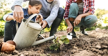 Kind trägt Gießkanne und bewässert Pflanze im Garten unterstützt von älteren Personen hält Schaufel in sonniger grüner Umgebung