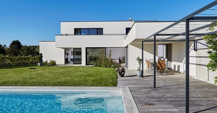 Galerie-Bild 5: Modernes weißes zweistöckiges Haus steht mit großen Fenstern und einem gepflegten Garten mit Terrasse. Ein Swimmingpool befindet sich im Vordergrund und blauer Himmel bildet den Hintergrund.