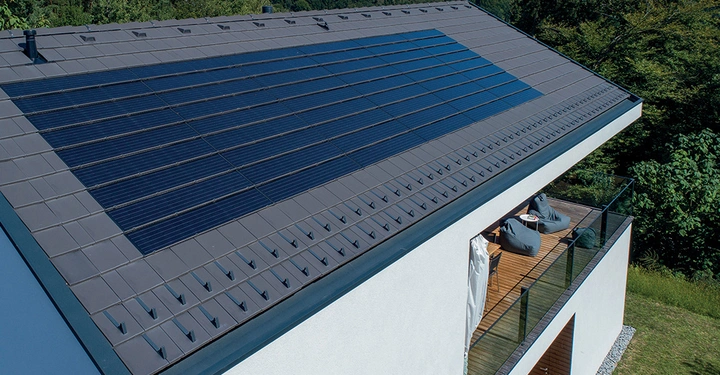 Solarmodule bedecken das Dach eines modernen weißen Hauses das sich in einer grünen bewaldeten Landschaft befindet. Auf der Terrasse sitzen Menschen und genießen die Aussicht.