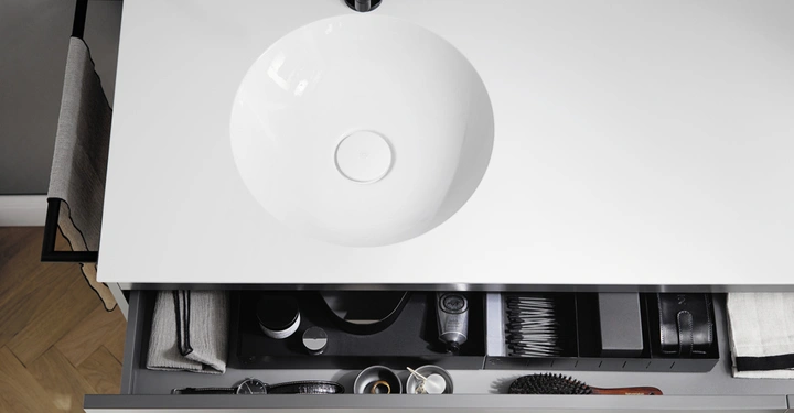 Weißes Waschbecken auf einer Arbeitsplatte mit geöffneter Schublade darunter in der organisierte Utensilien wie Handtücher Bürsten und Pflegeprodukte aufbewahrt sind. Heller Boden und Handtücher daneben.